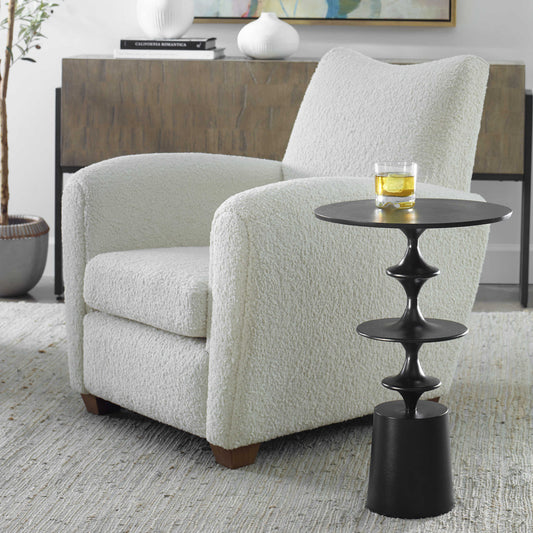 A contemporary accent table with a textured aluminum top and black satin legs, featuring a uniquely curved design, displayed in a living room setting with a chair and a glass of liquor on top.