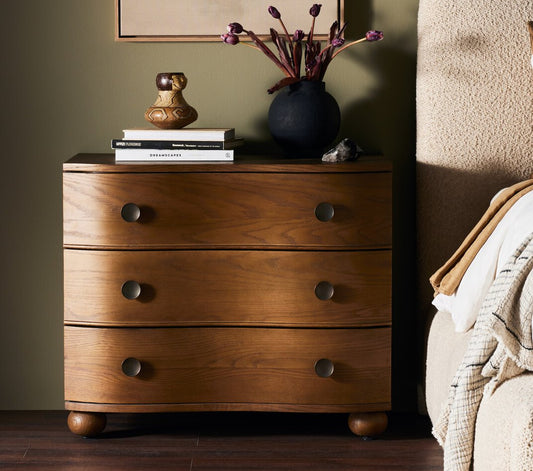 A toasted oak stain solid oak nightstand with three drawers and oversized hardware, placed in a bedroom setting.