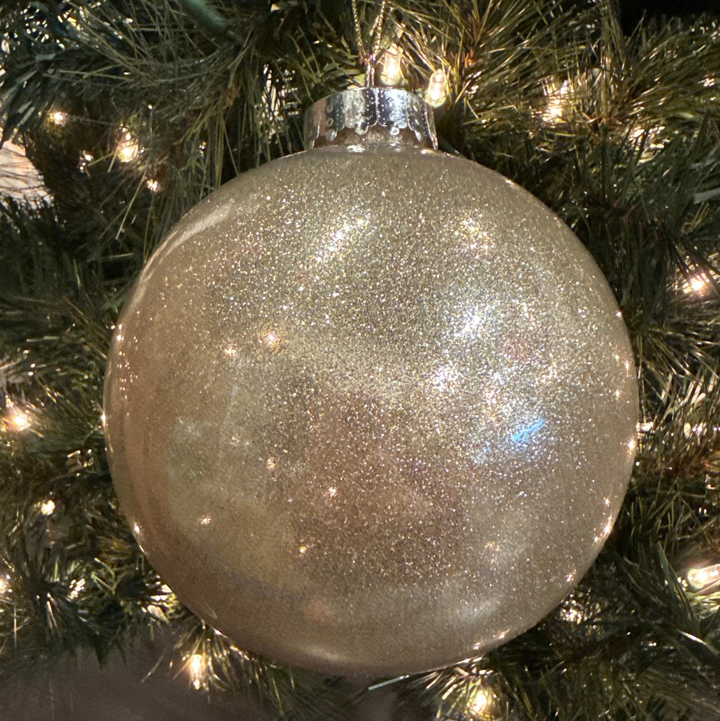 A clear spherical Christmas ornament with glitter and a silver cap attached to a Christmas tree.