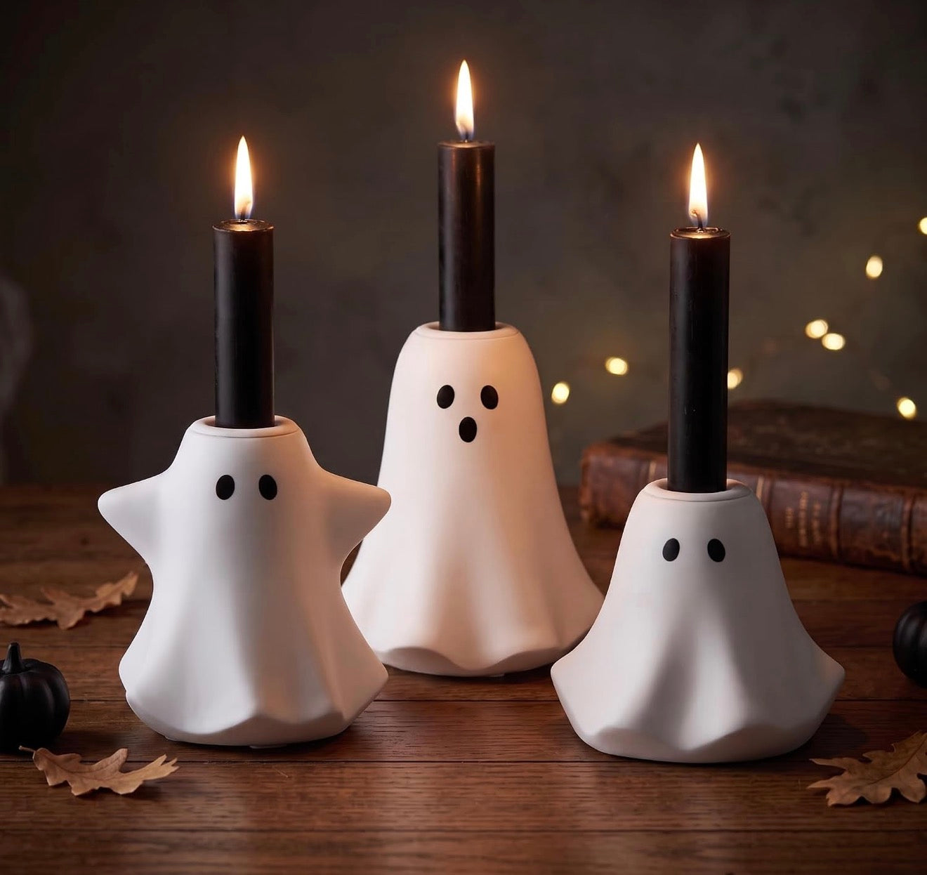 Three white ghost-shaped candle holders with black candle inserts, arranged on a wooden surface with autumn leaves and warm string lights in the background.