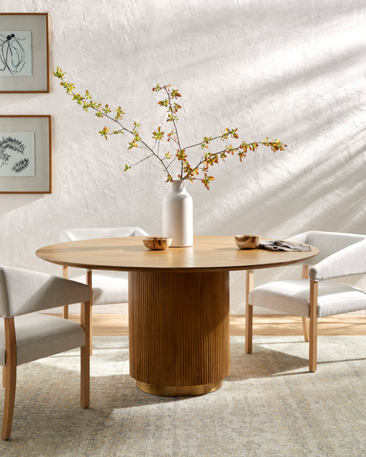 A 60" round dining table with a smooth top and reeded base, featuring brass detailing. The table is accompanied by a set of four chairs with a matching design in a room with natural lighting.