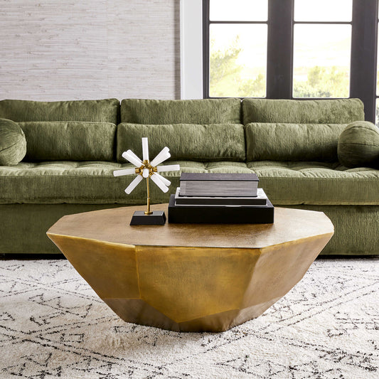 A gold-colored, faceted brass coffee table with a unique asymmetrical design, placed on a white area rug in a living room setting, with a green couch and a black bookshelf in the background.
