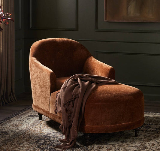 Rust velvet chaise with a blanket in a room with dark walls and a window.