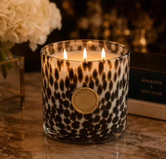 A lit candle in a clear glass jar with a black leopard print pattern and a round gold seal on the front, set on a marble surface with flowers and a book in the background.