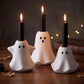 Three white ghost-shaped candle holders with black candle inserts, arranged on a wooden surface with autumn leaves and warm string lights in the background.