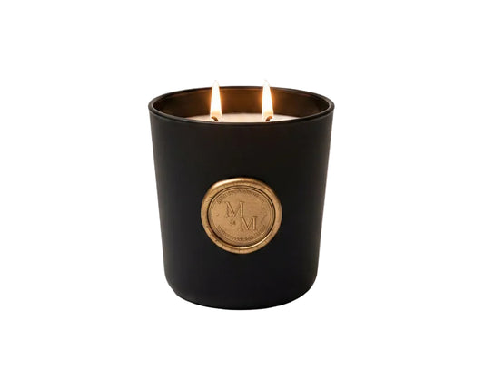 A black double-wick candle with gold detailing sits on a marble surface, styled with branded cards, books, flowers, and a glass vase in a warm indoor setting.