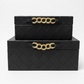 Two stacked black woven leather storage boxes with gold chain handles, displayed on a wooden table in a home decor store.