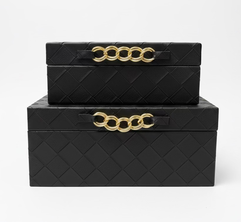Two stacked black woven leather storage boxes with gold chain handles, displayed on a wooden table in a home decor store.
