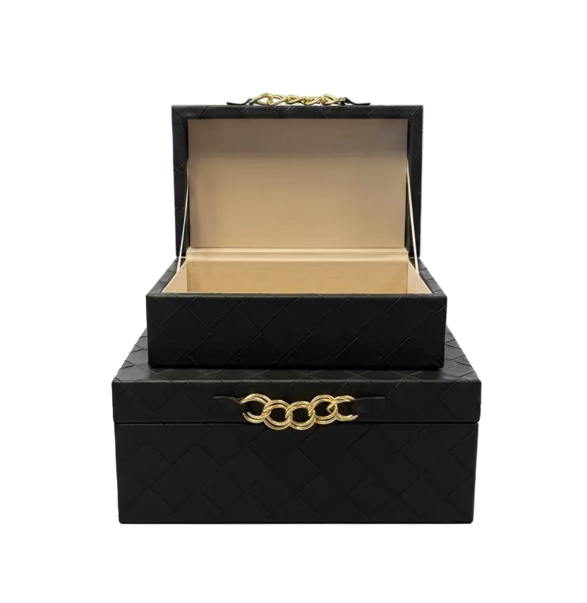 Two stacked black textured jewelry boxes on a wooden table, with the top box open to show a beige interior. The front box has a gold chain link handle, and the top box has a gold chain handle on the lid.