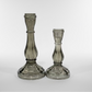 Two glass candle holders on a white surface with a pegboard background