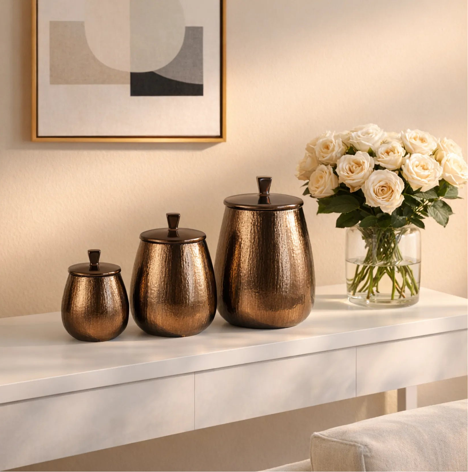 Decorative bronze canisters on a white surface with a vase of white flowers and abstract art in the background.
