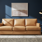 Brown leather sofa in a living room with a dark blue wall, abstract painting, and side table.