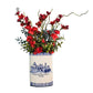 A ceramic vase with blue and white Portuguese tile detailing, filled with red artificial flowers and green leaves, displayed on a rustic wooden surface against a textured gray wall with decorative wooden lettering.