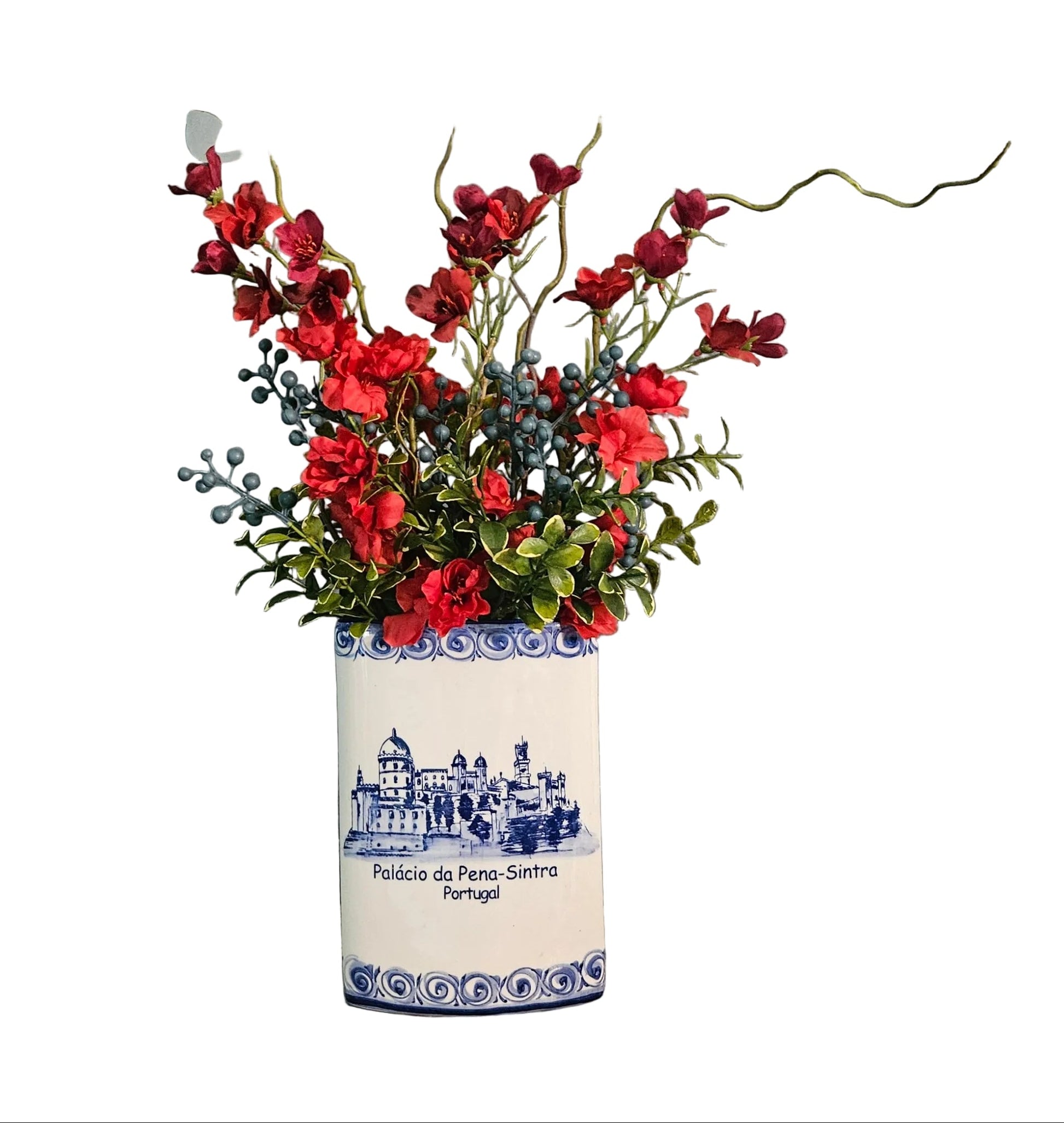 A ceramic vase with blue and white Portuguese tile detailing, filled with red artificial flowers and green leaves, displayed on a rustic wooden surface against a textured gray wall with decorative wooden lettering.