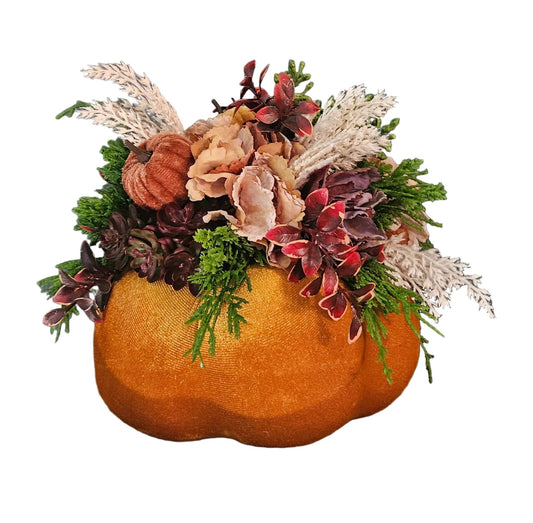 An autumn floral arrangement in a sculptural orange pumpkin-shaped planter, featuring faux ferns, dried grasses, and fall flowers, displayed on a rustic wooden surface against a dark gray wall.