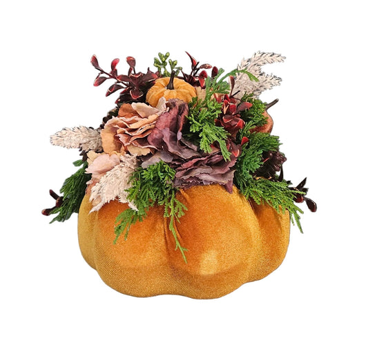 An autumn-themed floral arrangement in a small orange pumpkin-shaped pot, featuring green pine branches, muted pink and beige flowers, and burgundy accents, isolated on a white background.