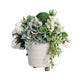 Faux succulent and floral arrangement in a white pot on a wooden surface with a gray wall background.